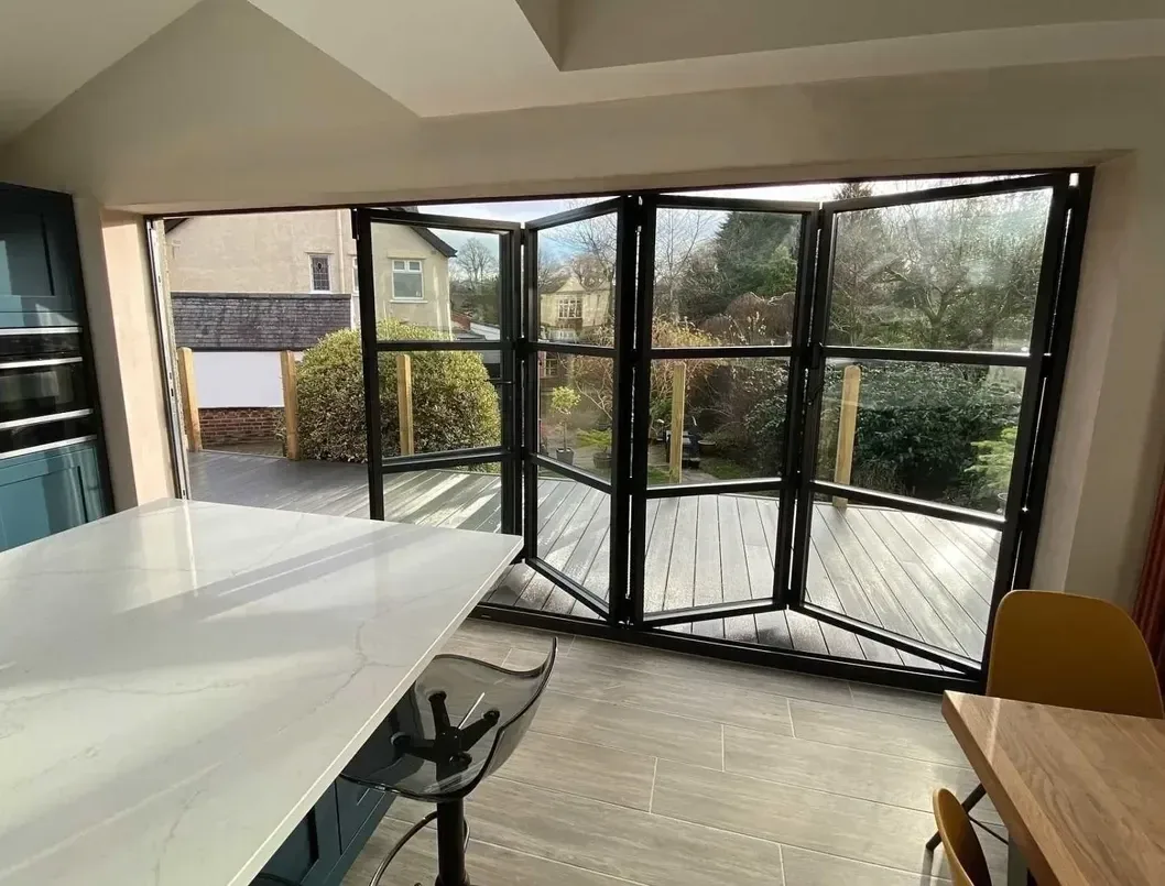 Black heritage-style aluminium bi-fold doors with glazing bars, folded open from a kitchen onto decking