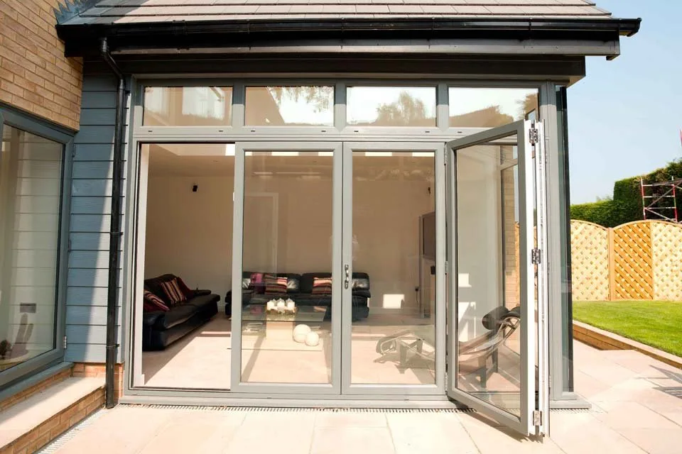 Grey aluminium bi-fold doors with glazed top lights, one panel open from a garden room onto a patio